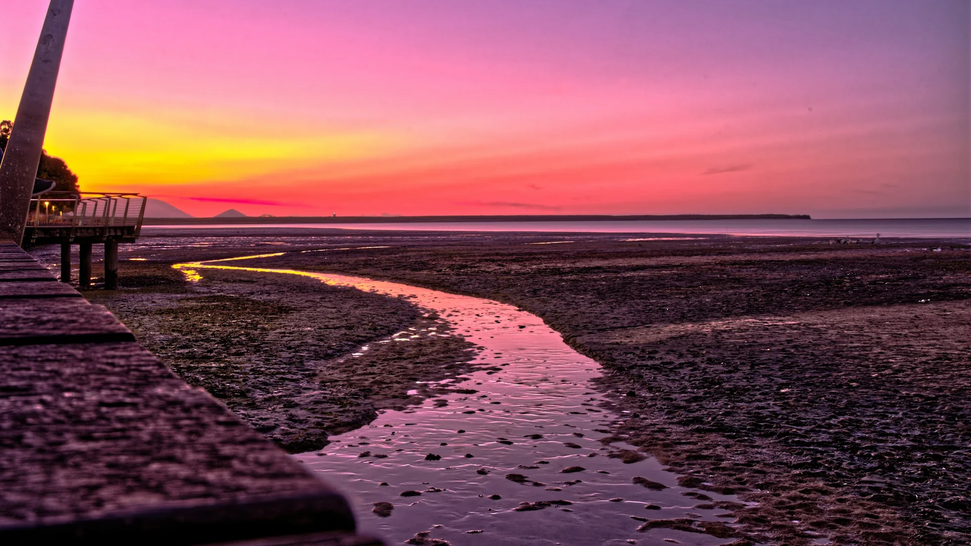 A purple, pink and gold sunset over the Cairns foreshore, with a still channel of water winding across the mudflats beside a timber boardwalk.