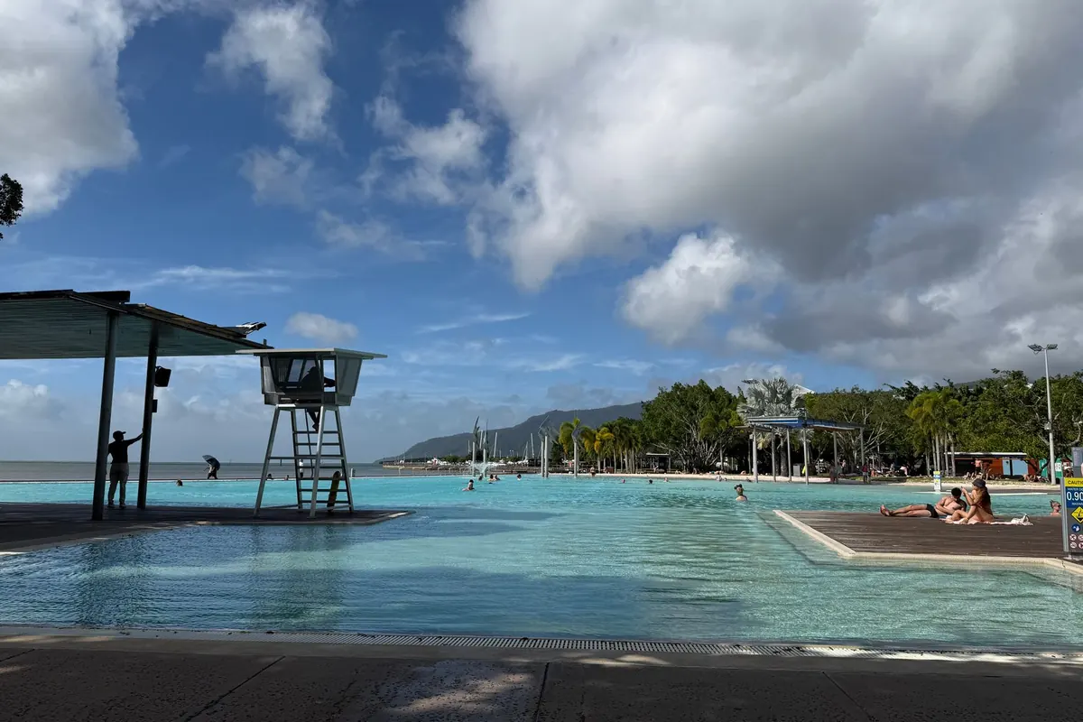 The Esplanade Lagoon in Cairns, with a lifeguard tower beside the pool, swimmers on a sun deck and green hills in the distance.