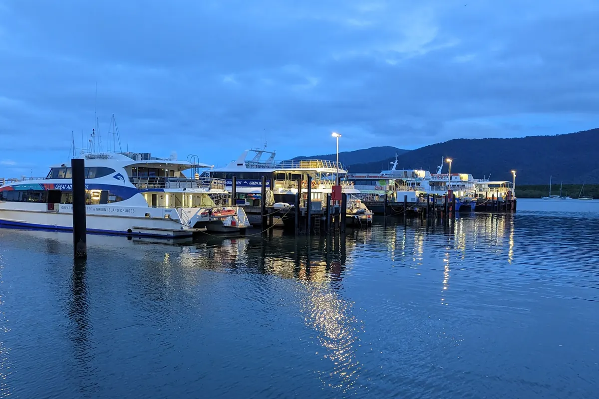 White and blue tour boats moored at a pontoon in Cairns at dusk, their lights reflected in still water, with hills behind