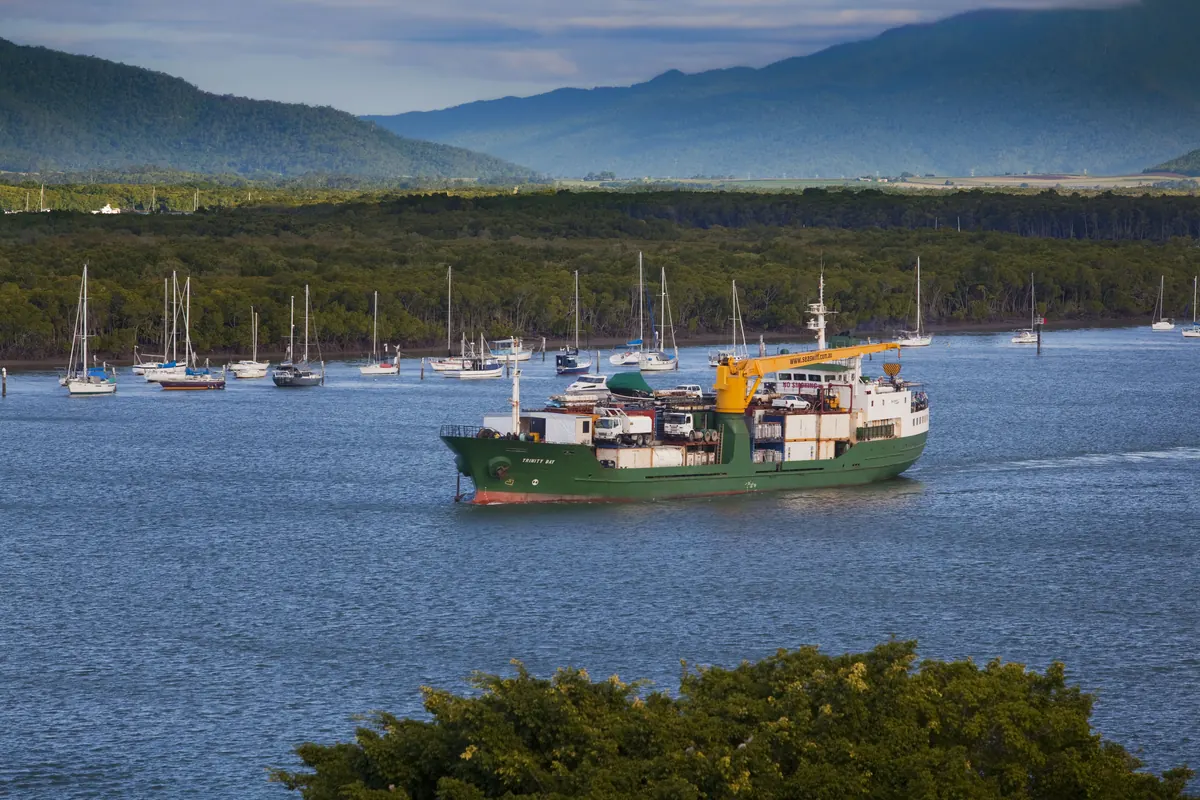 A green cargo ship with a yellow crane passing moored sailing boats on Trinity Inlet, with a dark line of mangroves and forested hills behind